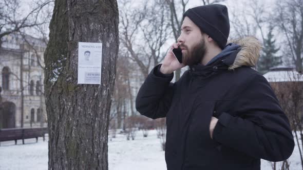 Portrait of Bearded Young Caucasian Man Standing Next To Missing Kid Ad and Calling on the Phone