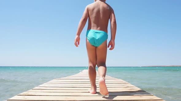 Happy Little Boy Dressed in Swimming Trunks Runs To the Azure Sea on a Wooden Pier on a Sunny Hot