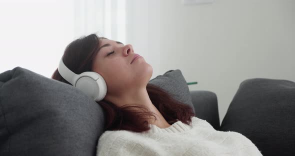 Portrait of Beautiful Woman Listening Music in Wireless Headphones While Relaxing on Grey Couch