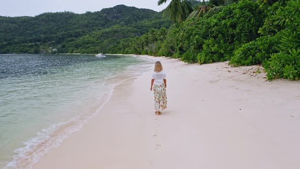 Camera Follows Young Woman As She Walk on the Paradise Sandy Beach with Green Foliage Palm Trees and