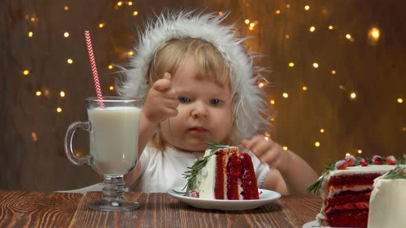 Llittle Girl in Santa Claus Hat is Sitting Near the Table with Red Velvet Cake