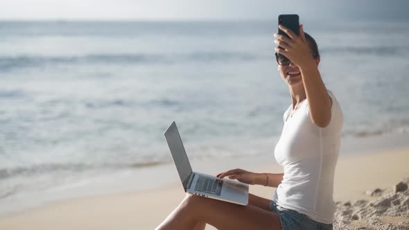 Woman with Laptop Sits on Beach Near Ocean and Uses Phone for Video Call