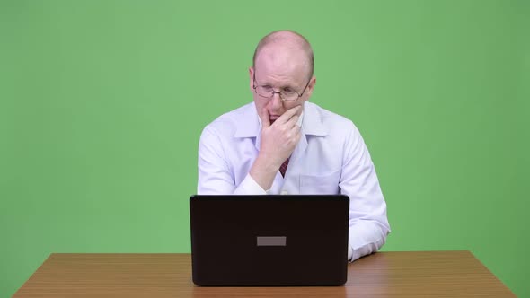 Mature Bald Man Doctor Thinking While Using Laptop Against Wooden Table