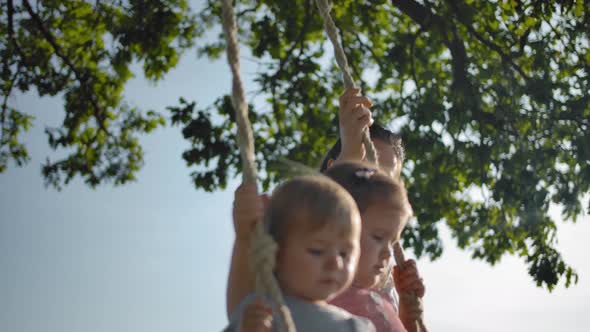 Mom Shakes Two Small Sisters on a Swing with Rope Handles