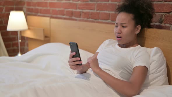 African Woman Reacting to Loss on Smartphone in Bed