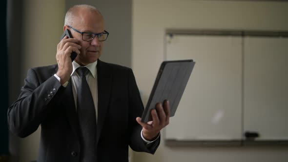 Senior Businessman Talking On The Phone While Using Digital Tablet By The Window At Work
