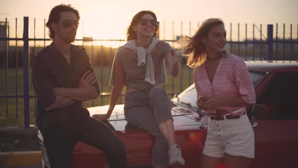 Friends Sit on the Hood of a Car Talk and Laugh at Sunset