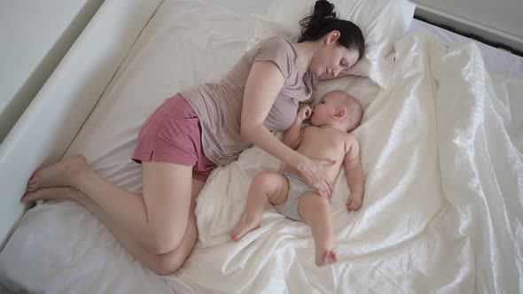 Tired Mother Lying with Newborn Cute Infant Hyperactive Baby Boy on Bed Holding Him on Arms Hugging