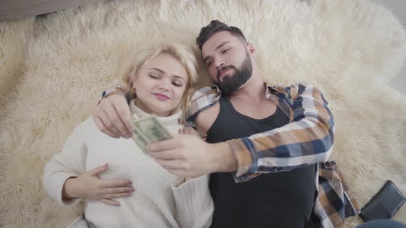 Happy Rich Caucasian Couple Lying on Soft Carpet and Counting Dollars. Blond Girl in White Sweater