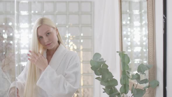 Young Caucasian Woman With Long Blond Hair Wearing a Bath Towel Looking at Camera and Combing