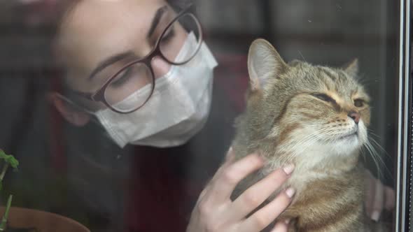 A Young Woman in a Mask Strokes a Cat. The Girl in Quarantine at Home Stands at the Window and