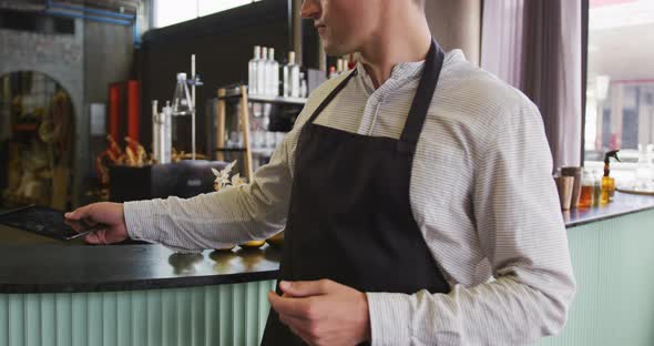 Portrait of caucasian man wearing apron working at a bar, using tablet and smiling to camera