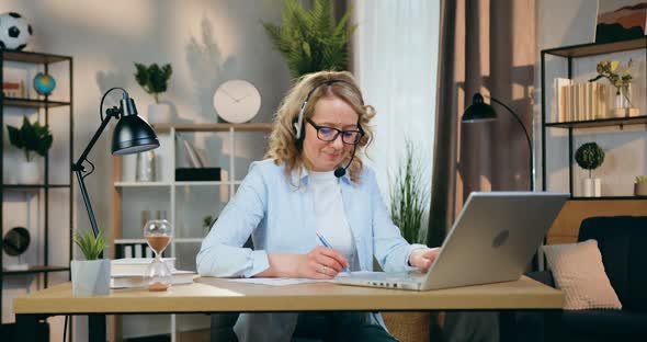 Woman in Headset Working with Documents and Information on Laptop at Workplace in Cozy Room