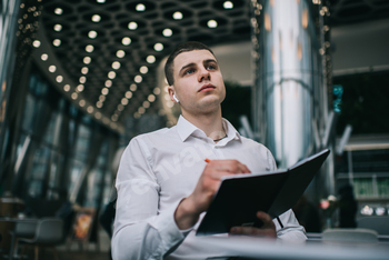 Young male entrepreneur in wireless earbuds thinking and taking notes