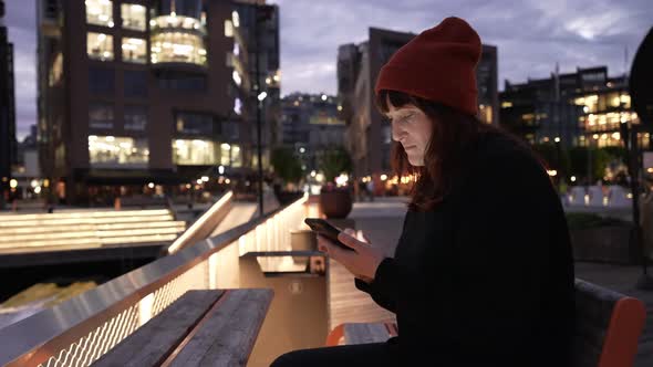 Young Hipster Girl Communicates on the Internet Using a Modern Smartphone on the Street at Night