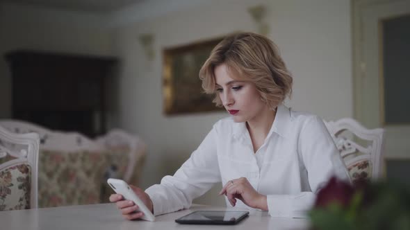 Happy Woman Uses Smartphone and Tablet at Table in Light Room