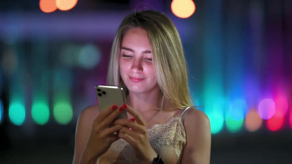 Cute Girl Uses Smartphone and Smiles Standing on Night Street in the City