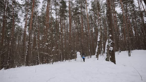 Boy in Jacket Hat Runs Around Snowman in Snowy Forest