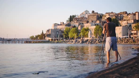 A Man Wearing Shorts Standing In The Lake Bracciano Shore With His Camera Capturing The Beauty Of Ca