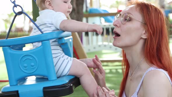 Redhaired Woman Swinging Her Young Son on Plastic Swings