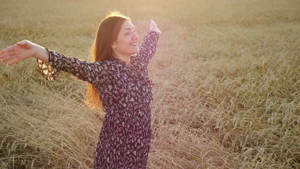 Young Woman in Dress Smiling and Raises Her Hands Up While Standing in a Field of Ripe Wheat