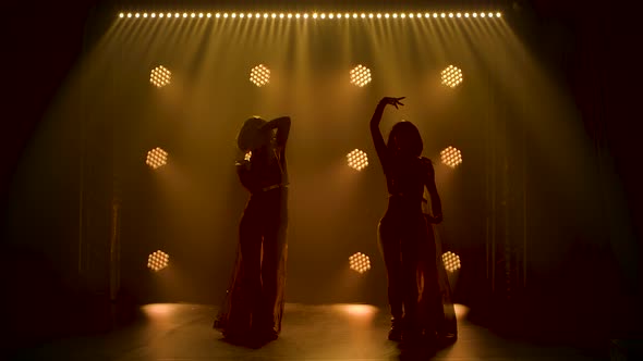 Silhouettes of Two Women Dancing and Waving Their Capes in a Dark Smoky Studio with Soft Yellow