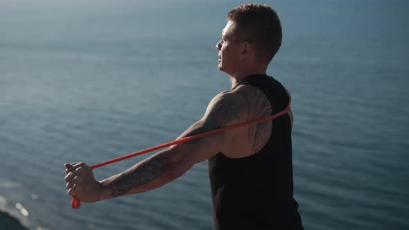 Muscular Man with Tattoo Trains with Fitness Elastic Bands Outdoor with Sea View