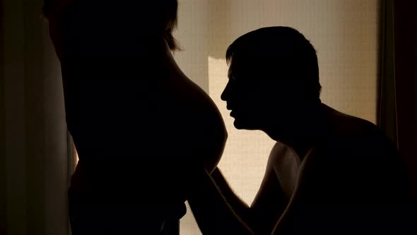 Husband Kisses Pregnant Wife Tummy Silhouette Against Window And Roll Curtains