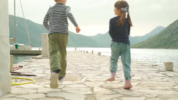 A Boy and a Girl Run Along the Pier Against the Backdrop of the Sea and Mountains