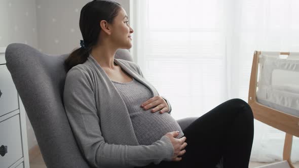 Zoom in of caucasian woman in advanced pregnancy sitting on armchair and stroking the abdomen. Shot