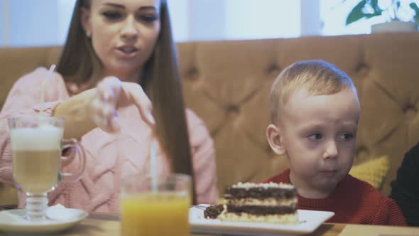 Little Boy Eats Cake and Talks to Mother at Wooden Table