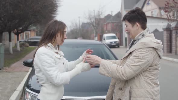 Side View of Caucasian Mature Senior Woman Giving Car Keys To Young Brunette Girl in Front