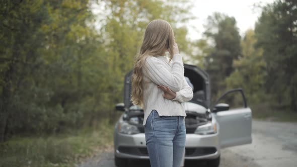 Charming Slim Young Woman Talking on the Phone As Man Repairing Car Engine at the Background