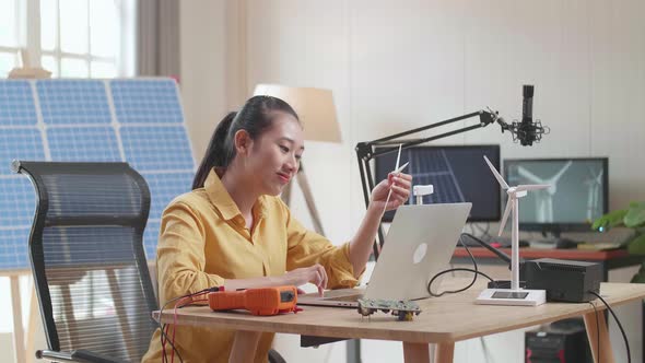 Asian Woman Looks At Wind Turbine While Working With Laptop Next To The Solar Cell At The Office