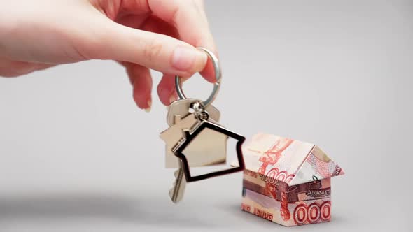 A woman's hand shakes keys with a keychain next to an origami house made of Russian rubles.