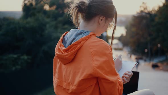 Young and Hardworking Woman Makes Notes Positioned on a Bench in the Park