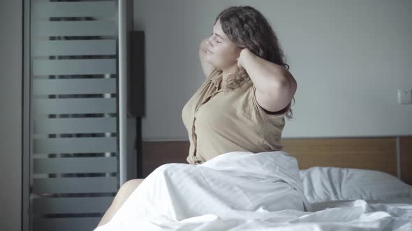 Side View of Rested Young Caucasian Plump Woman Sitting on Bed in the Morning and Stretching