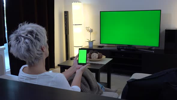 A Middle-aged Woman Sits, Watches TV with Green Screen and Looks at a Smartphone with a Green Screen