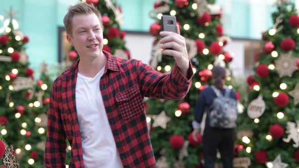 Happy Young Man Using Phone And Video Calling Against Christmas Trees Outdoors