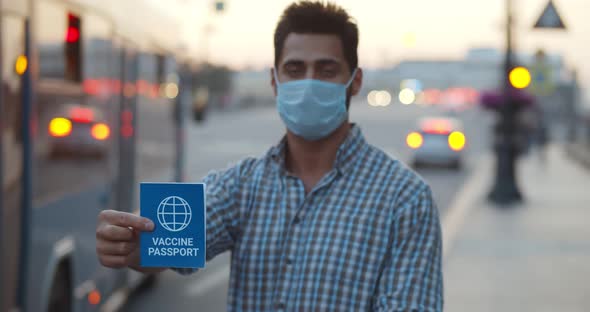 Medium Shot of Man in Mask Looking at Camera Holding Vaccine Passport Outdoors