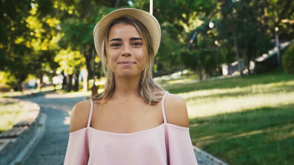 Young Female in Hat and Pink Dress is Looking at you and Smiling Posing in Park