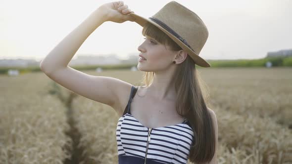 Portrait of Flirting Girl in Hat Poses and Smiles at Camera in Wheat Field