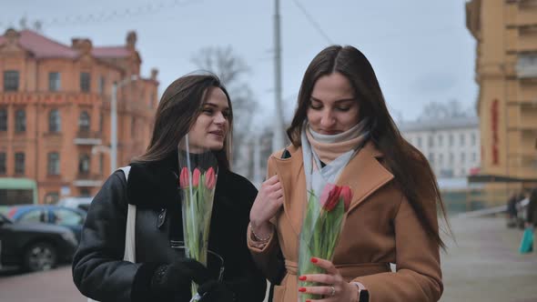 Two Joyful Friends in a Jacket and Coat are Walking Around the City with Tulips