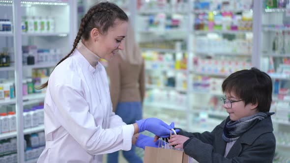 Smiling Woman Passing Shopping Bags with Medications to Boy in Drugstore Waving Goodbye to Client