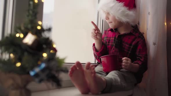 cute boy in santa hat t sitting on window sill drinking milk