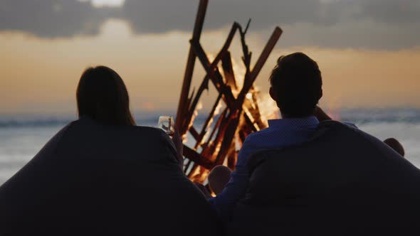 Happy Couple Resting in Front of Burning Yellow Campfire at Evening on the Coastline