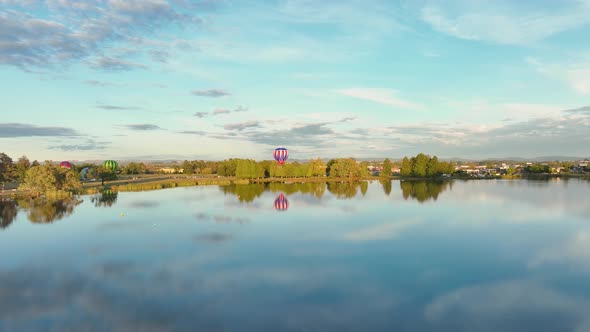 Hot air balloon rising through low trees and emerging to cross lake and into city.