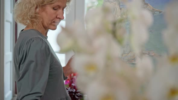 Middleaged European Woman Artist Paints a Picture on an Easel with Acrylics in Front of the Window