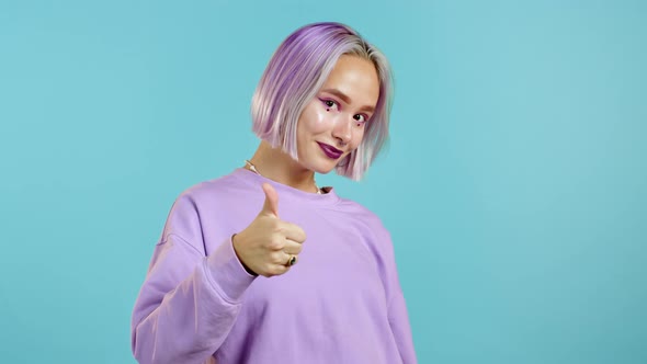 Cute Woman with Violet Dyed Hairstyle Showing Thumb Up Sign Over Blue Background. Positive Young