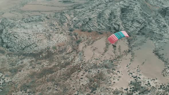 Aerial View Of Flying Motorised Paraglider Flying Over Dry Rocky Desert Landscape. Follow Shot